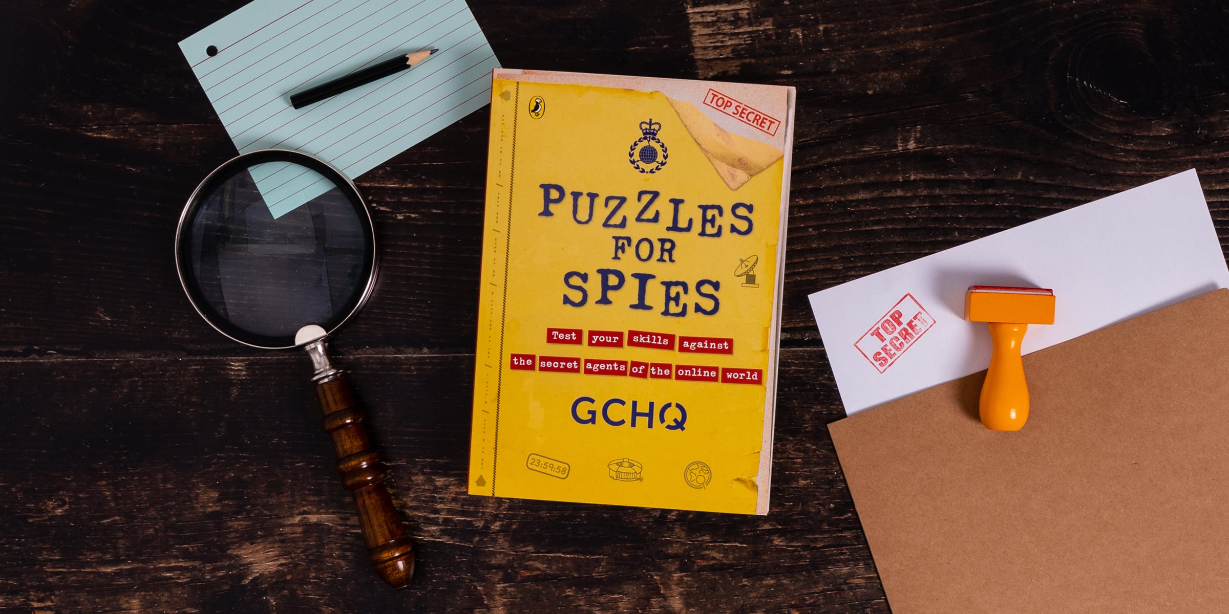 Puzzles for Spies book on a desk with a top secret document, a magnifying glass on a dark table