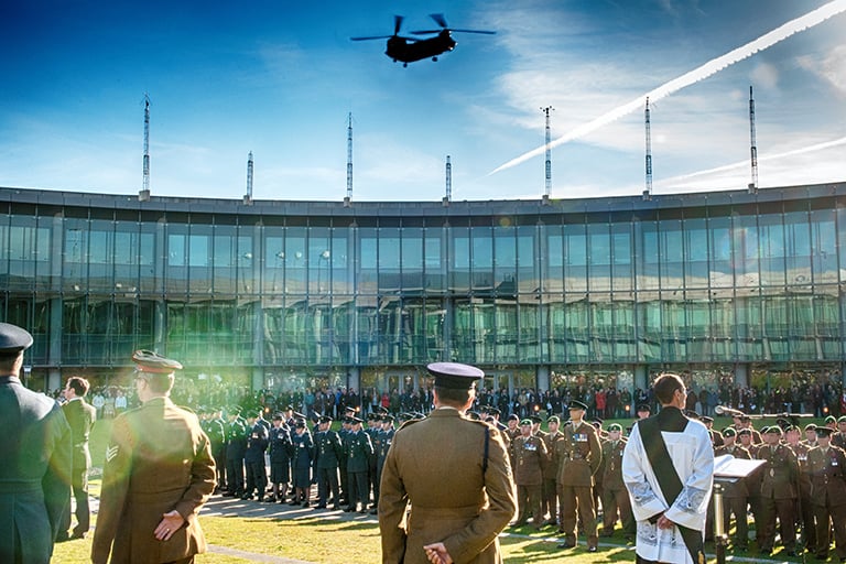 People in standing during the service, military and civilians. A large helicopter in the sky above