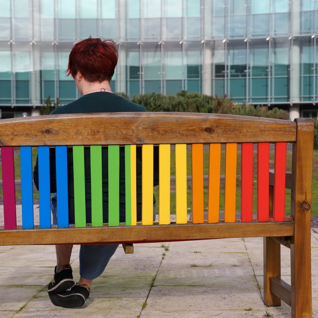 Jane is sat on a bench with her back to the camera. The bench is painted in the colours of the Pride flag.