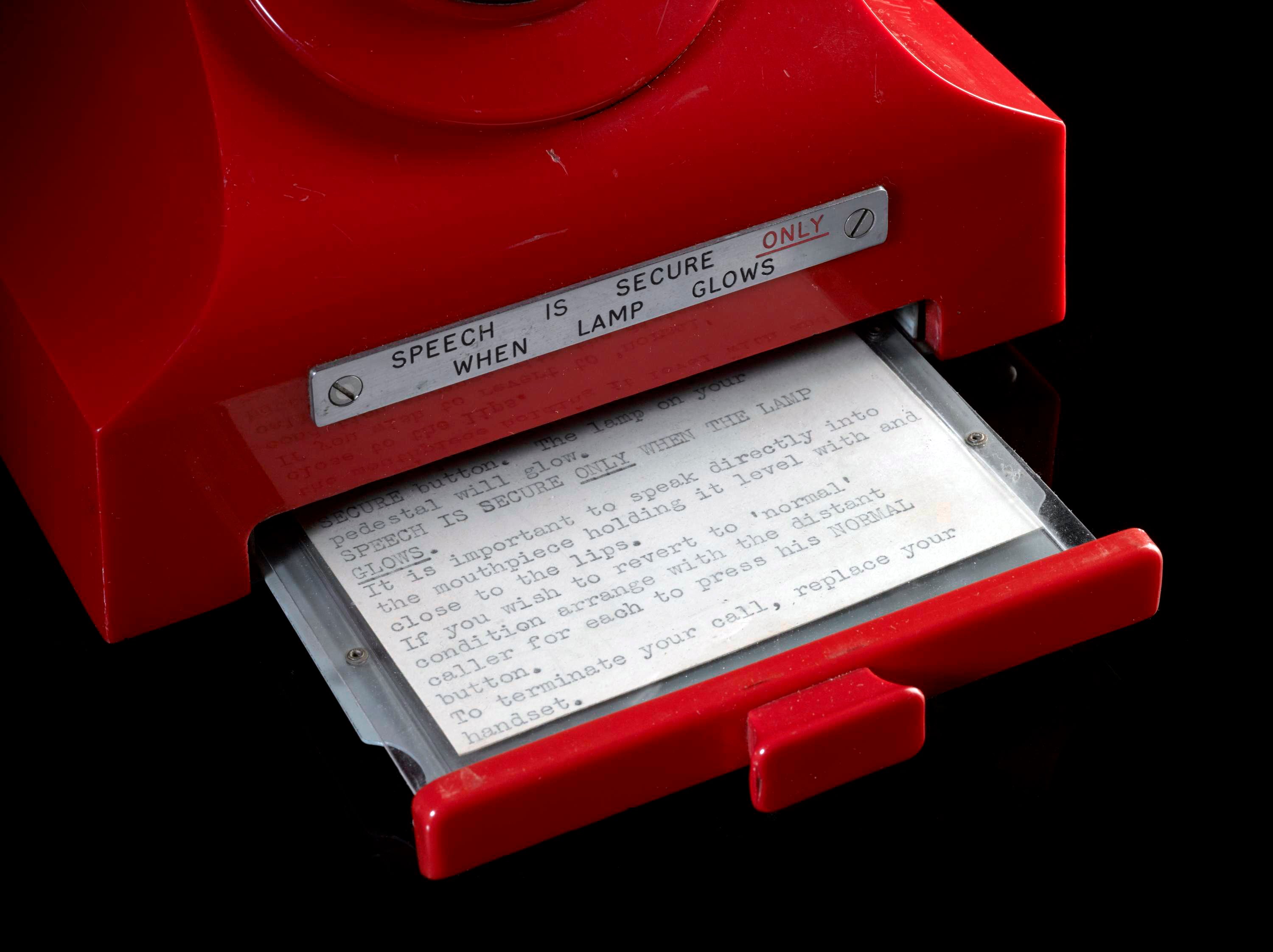 Close-up of an old-fashioned red telephone, showing its instruction label which reads: speech is secure only when the lamp glows