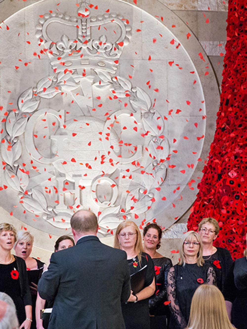 A choir sings in front of the GCHQ poppyfall in the atrium as poppies fall from the sky.