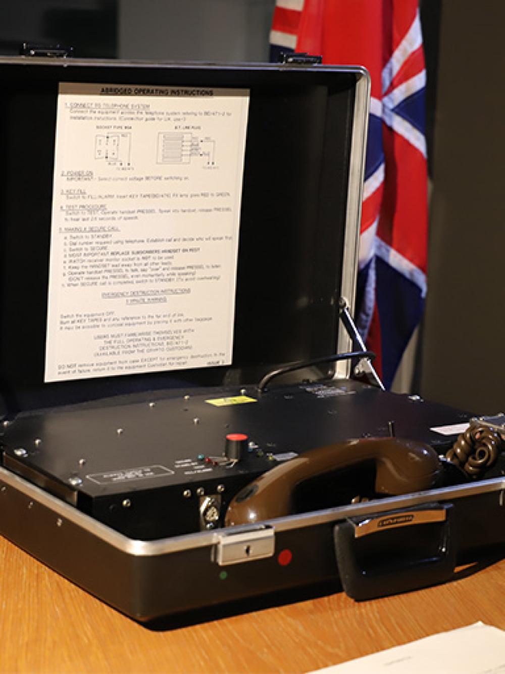 The Brahms secure portable telephone housed within an open briefcase on a desk with the Union Jack in the background
