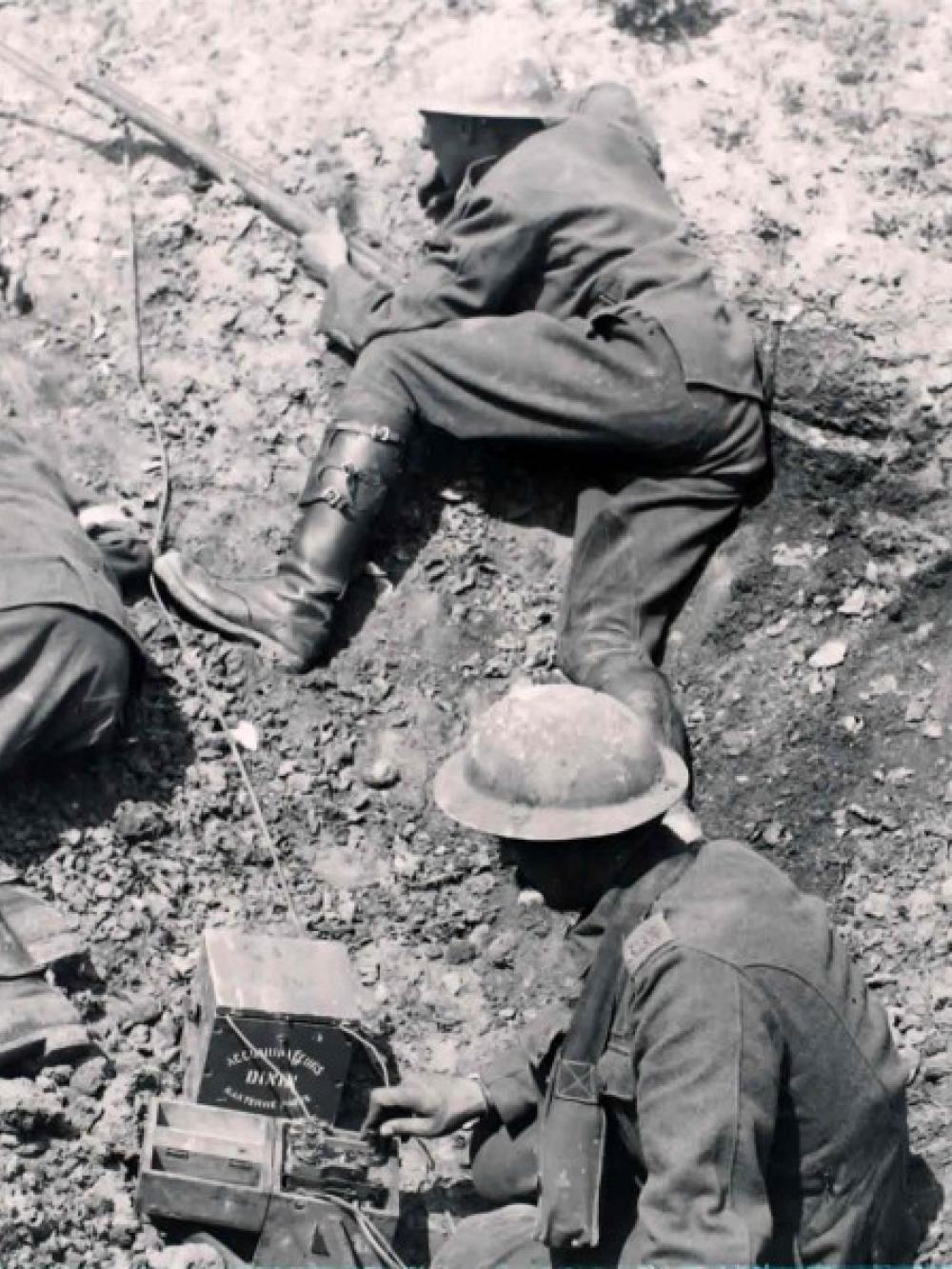 Black and white photo of soldiers in a large depression in the ground. One soldier is pressing a lever on a device in a wooden box to generate morse code. Wires are seen leading into the depression and connected to the box.