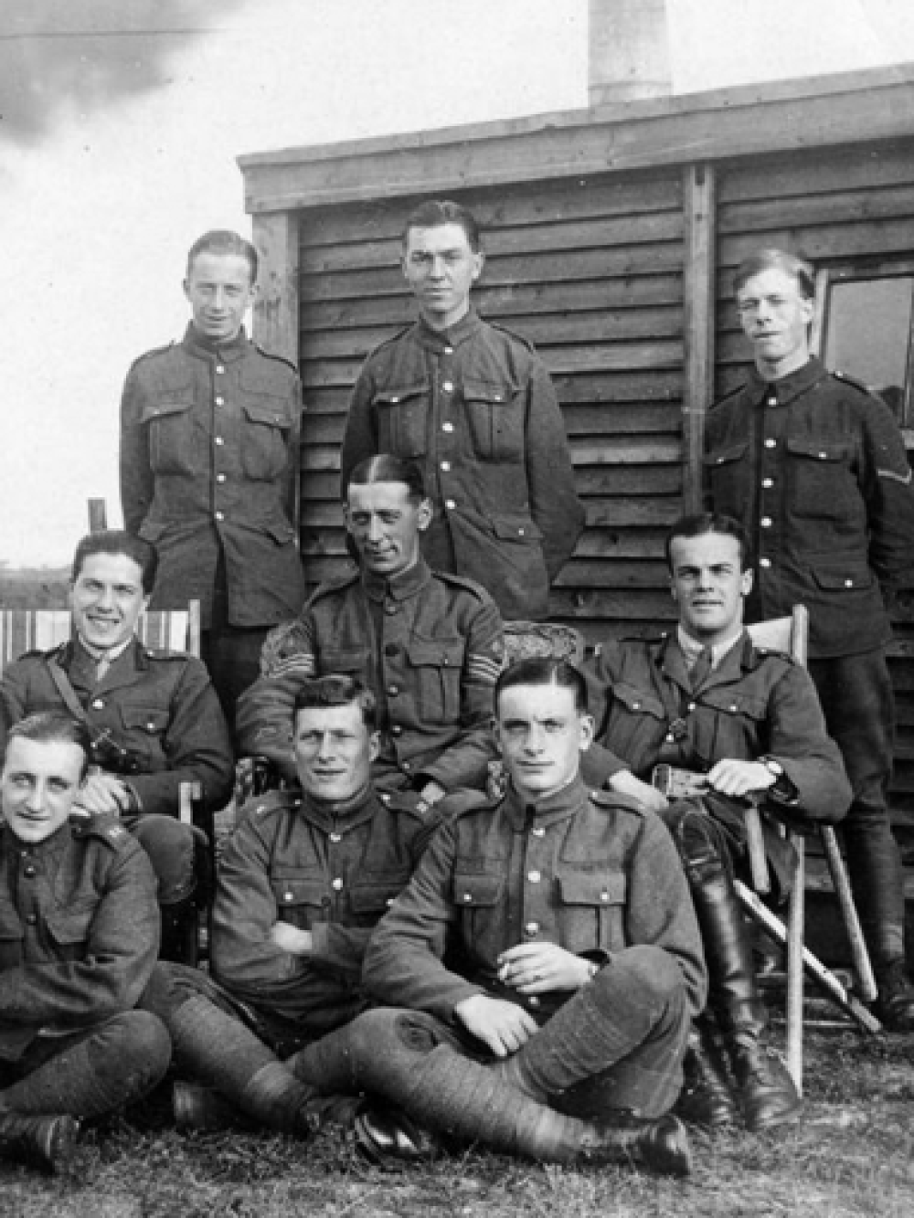 9 men in WWI uniform posing in front of a large shed and field
