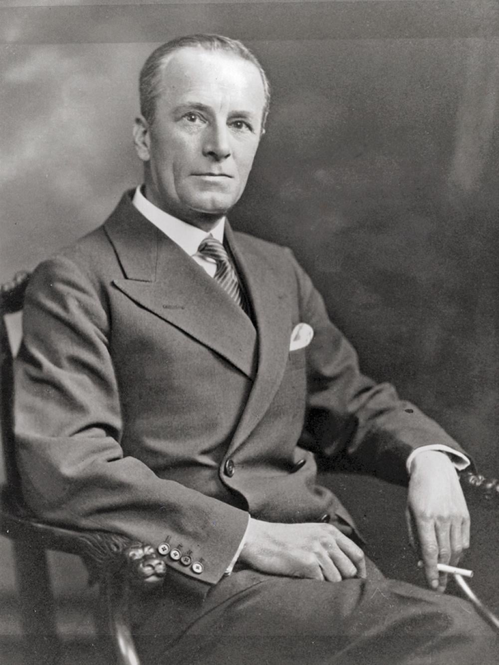 He is wearing a suit and tie, sat on a carved chair and holding a cigarette