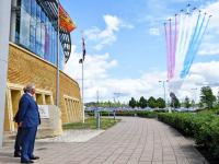 Ten Red Arrows (RAF Hawk T1As) in a V formation flying over GCHQ, producing red, white, and blue comtrails, while HRH Prince Charles looks on.