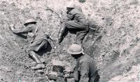 Black and white photo of soldiers in a large depression in the ground. One soldier is pressing a lever on a device in a wooden box to generate morse code. Wires are seen leading into the depression and connected to the box.