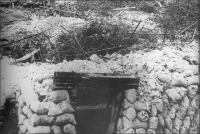 Wooden door frame and lintel with sandbags either side leading downwards. Behind lies the bare soil battleground covered with barbed wire and metal frames to stop infantry.