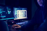 A hooded hacker working at a laptop in the dark with other computer screens behind him.