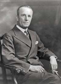 He is wearing a suit and tie, sat on a carved chair and holding a cigarette